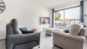 A modern living room with a grey sofa, a beige sofa, and a small round table. There is a large window with curtains, a flat-screen TV mounted on the wall, and a view of the outdoors. - 125m² Villa with Heated Pool, 3 Bedrooms, Terrace in La Flotte (La Flotte)