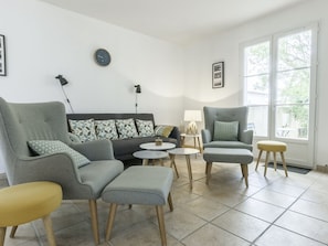 A living room with a grey sofa, two armchairs, a small round table, and a yellow stool. There is a window with a view of trees and a door. - Single-story house with enclosed garden for 6 people (Sainte-Marie-de-Ré)