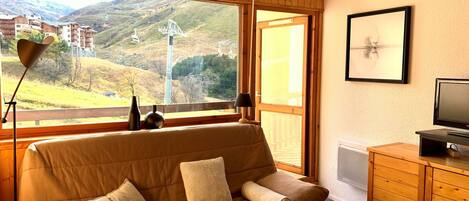 A cozy room with a sofa, a television, and a wooden cabinet. A large window offers a view of a mountainous landscape.