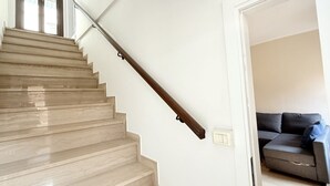 A staircase with a wooden handrail, a light-colored wall, and a gray sofa in the adjacent room. - O03 – Osimo, One-Bedroom Apartment with Terrace and Wi-Fi (Osimo)