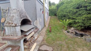 A rustic outdoor setting with a stone-built wood-fired oven, a ladder, and a small garden area with a pond and greenery. - Ferien_Haus & Groß Grundstück in Bad Elster (Bad Elster)