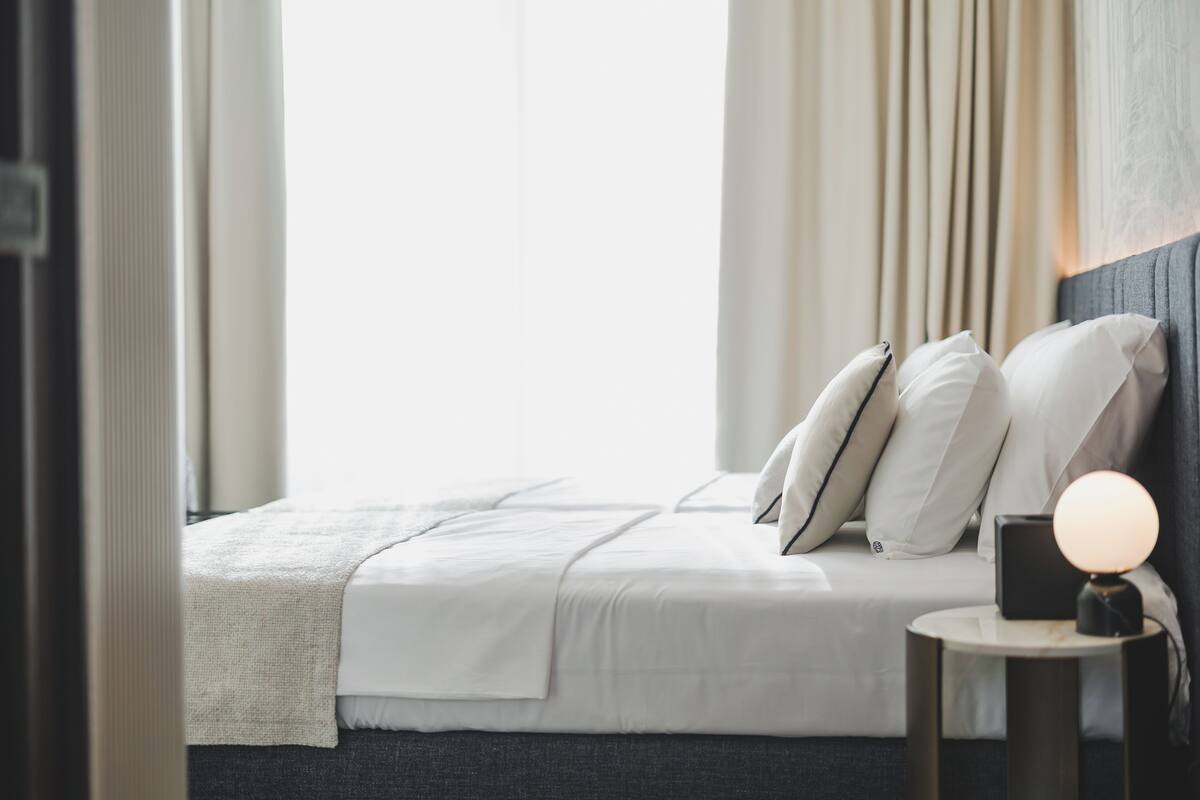 Un lit bien fait, avec du linge de lit blanc et des oreillers, une table de chevet avec une lampe, et une vue sur une fenêtre avec des rideaux.