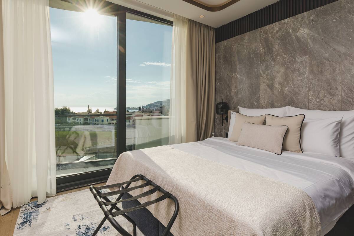 Une chambre moderne avec un grand lit, une vue sur la ville à travers une porte vitrée et un panorama urbain visible à l’extérieur.