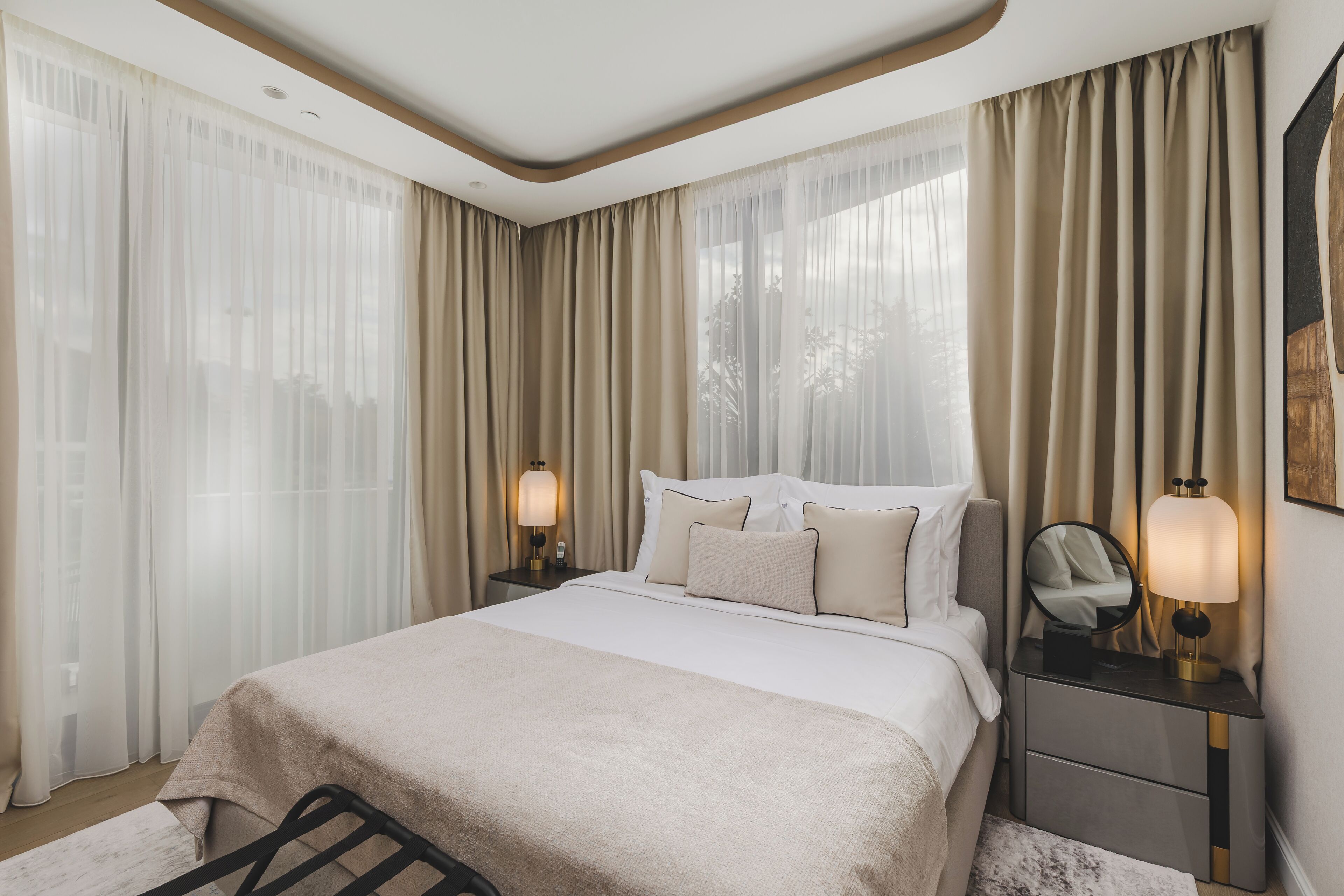 A bedroom with a bed, two bedside tables with lamps, a mirror, and sheer curtains.