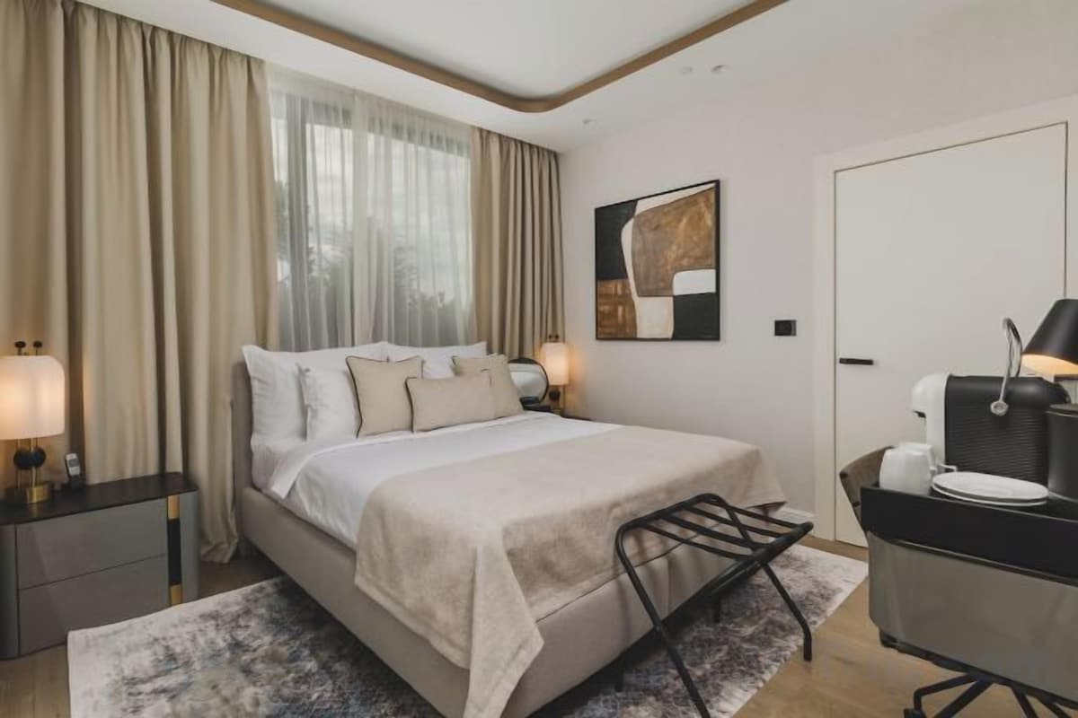 Habitación de hotel con cama, lámparas de noche, escritorio con silla, ventana con cortinas y una puerta.