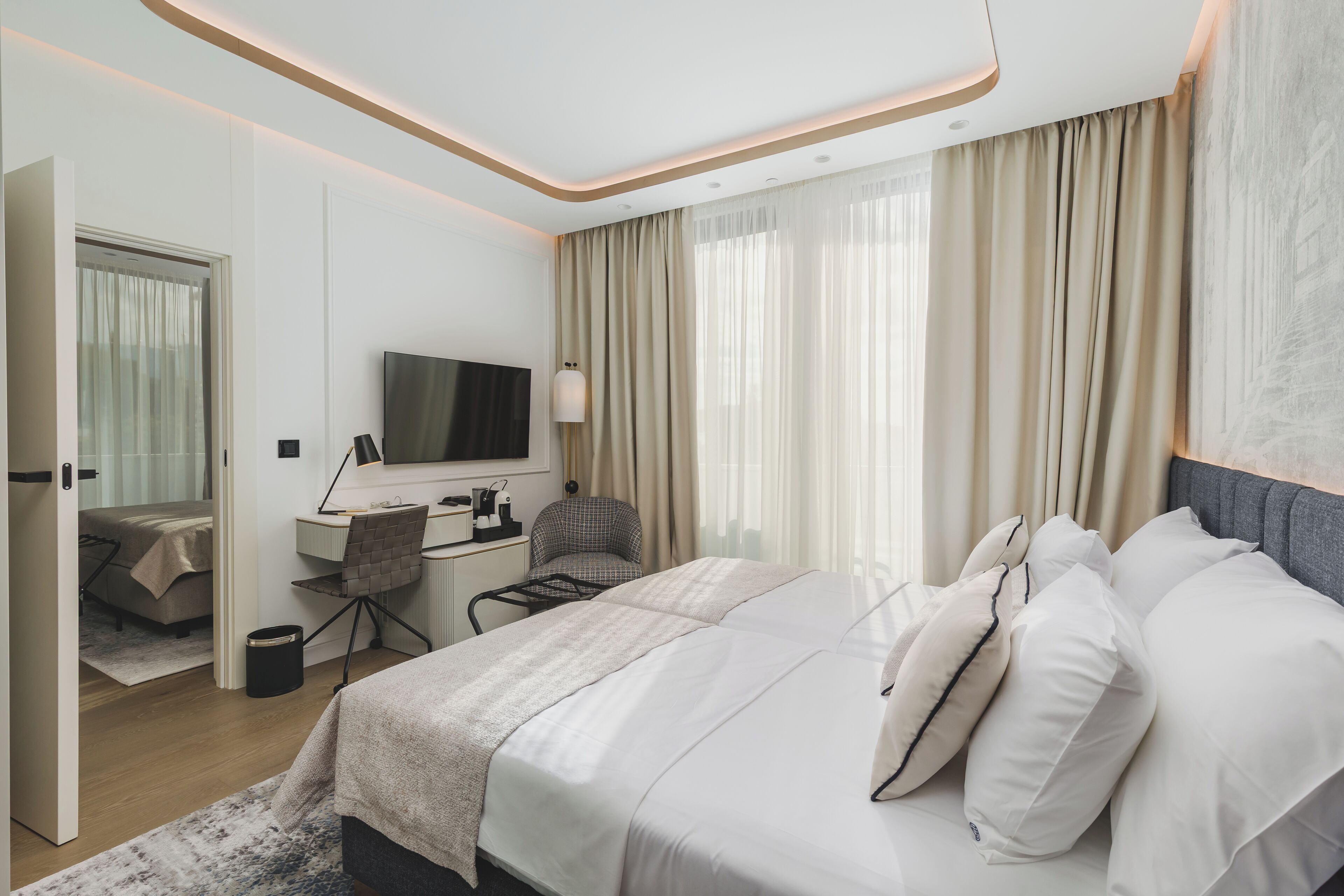 Una habitación de hotel moderna con una cama grande, un escritorio con silla, televisión y vistas a la ciudad a través de amplias ventanas.