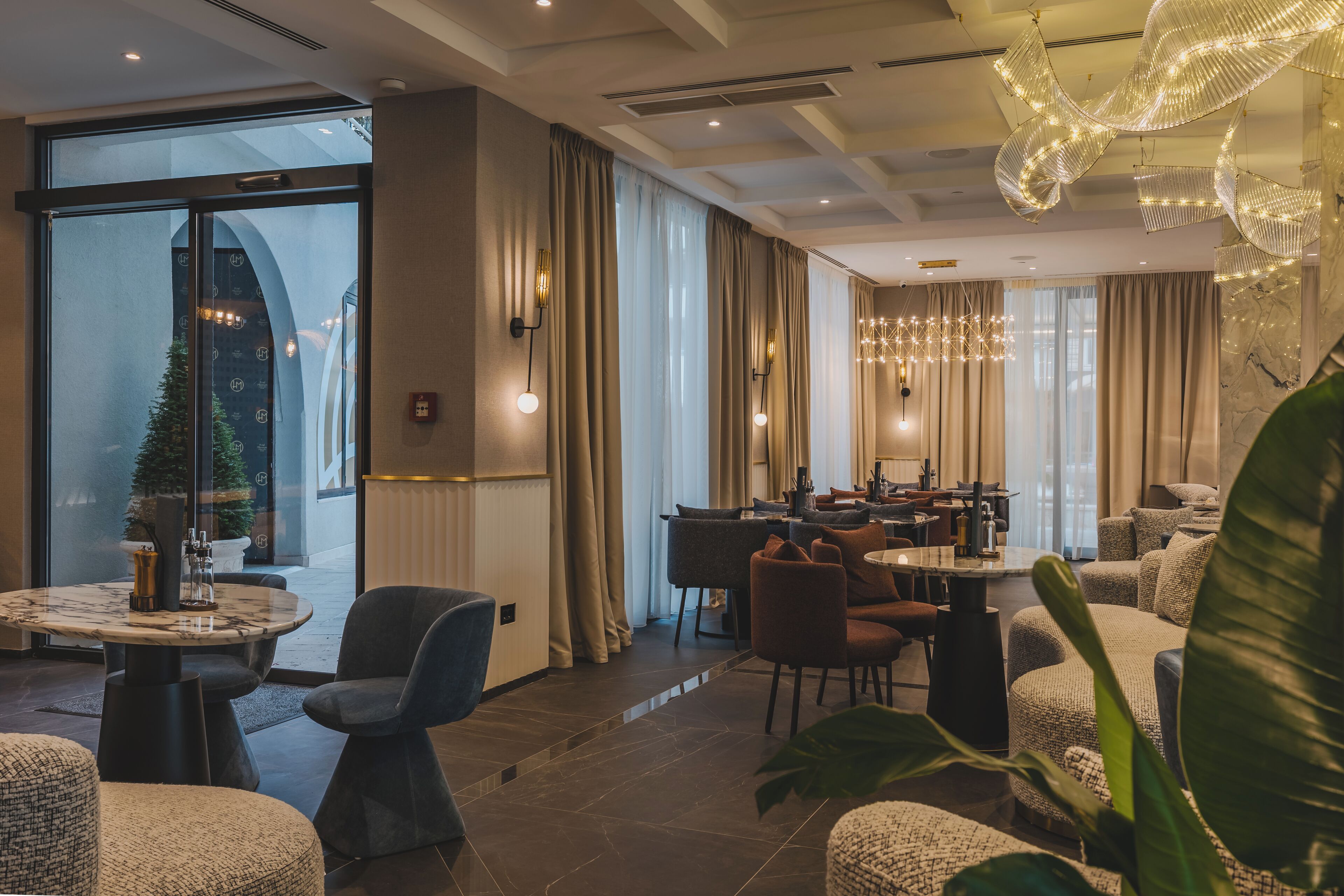 A modern hotel lobby with a dining area, large windows, and a decorative chandelier.