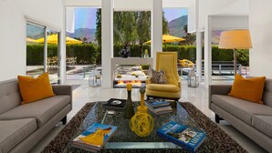 A modern living room with a glass coffee table, two sofas with yellow cushions, a yellow armchair, and a view of a pool and mountains.