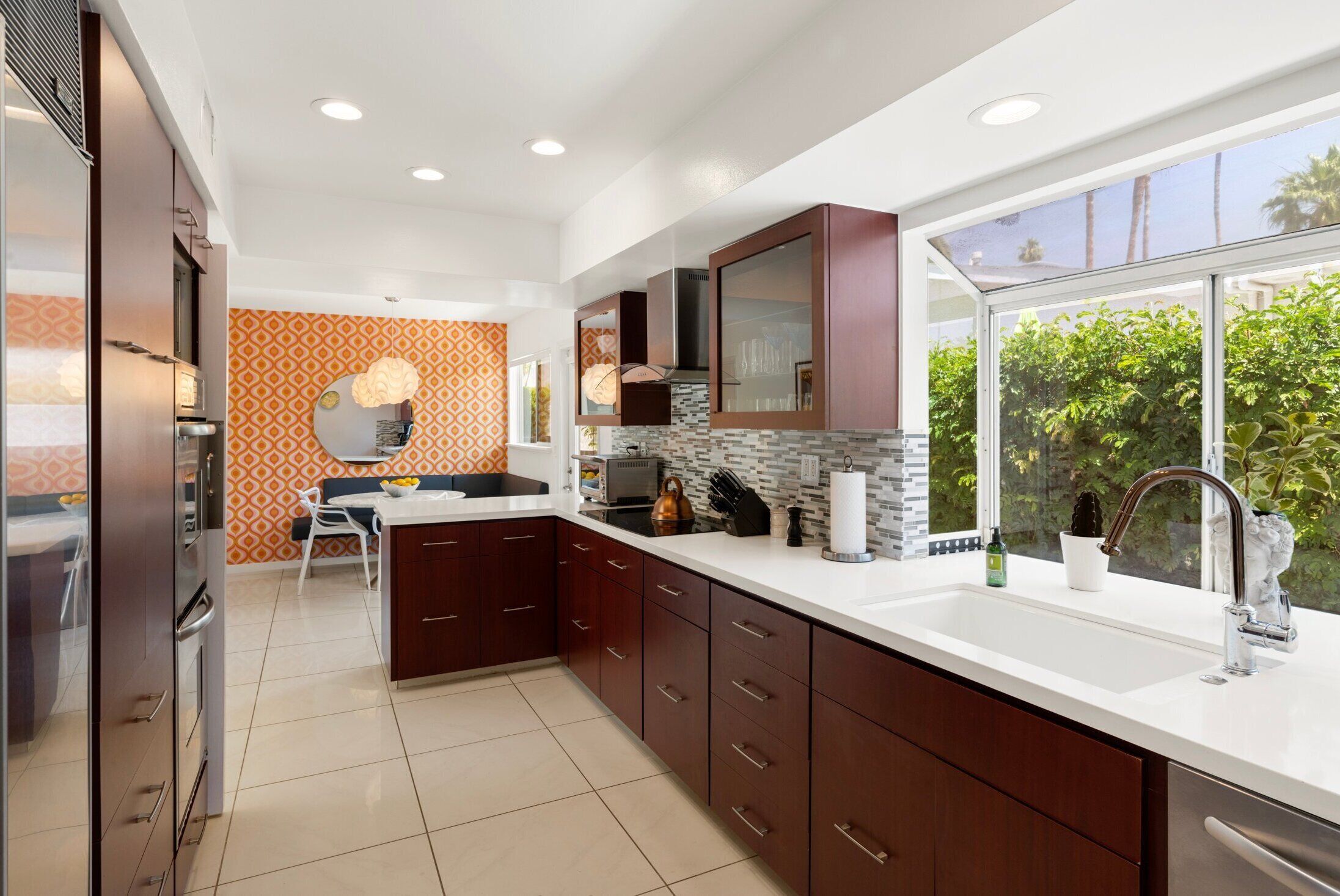 A modern kitchen with dark wooden cabinets, a white countertop, and a large window offering a view of greenery.