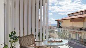 A balcony with a wicker chair and a small table, a potted plant, and a view of buildings and the sky. - Olenia Suites (Neos Marmaras)