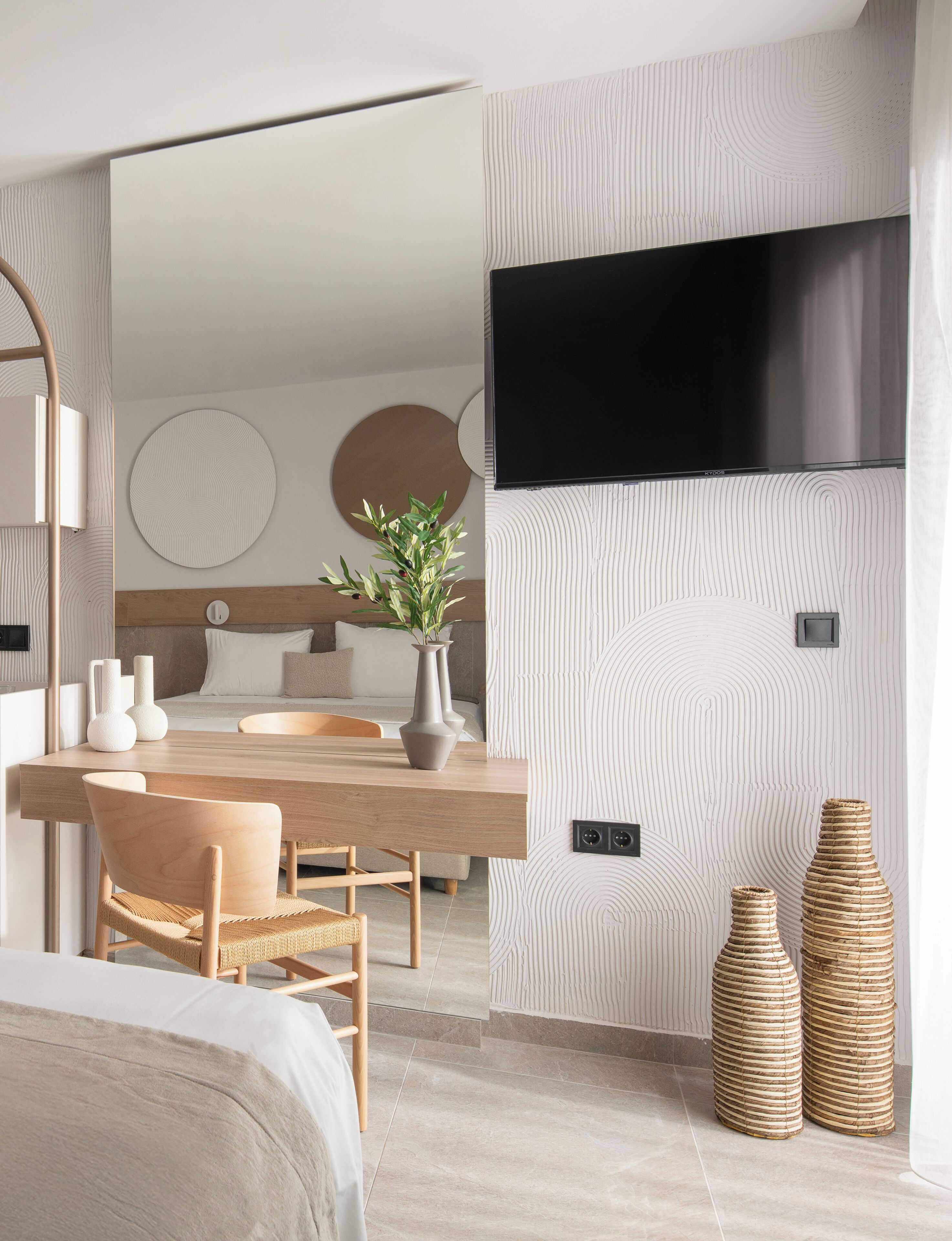 Una habitación de hotel moderna con una mesa de comedor de madera, un sofá, un televisor montado en la pared y un espejo grande.