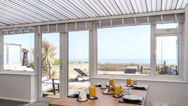 A sunlit dining area with a table set for breakfast, overlooking a beach and ocean view. - Beach Cottage with garden that blends into the beach in Pevensey Bay (Pevensey Bay)
