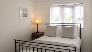 A bedroom with a bed, a nightstand, a lamp, and a window with blinds. - Beach Cottage with garden that blends into the beach in Pevensey Bay (Pevensey Bay)