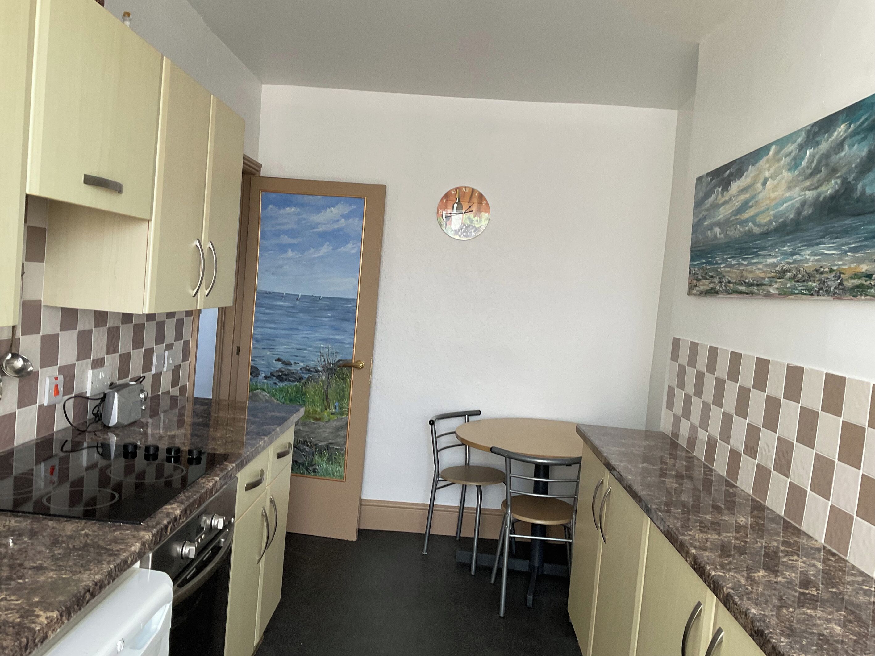 A compact kitchen with a stove, sink, and a small dining table with chairs. A door leads to a view of the sea.