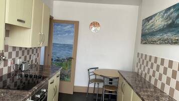 A compact kitchen with a stove, sink, and a small dining table with chairs. A door leads to a view of the sea.