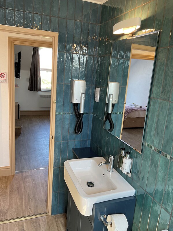 A bathroom with a white sink, two wall-mounted hair dryers, a mirror, and a shower area visible through an open door.