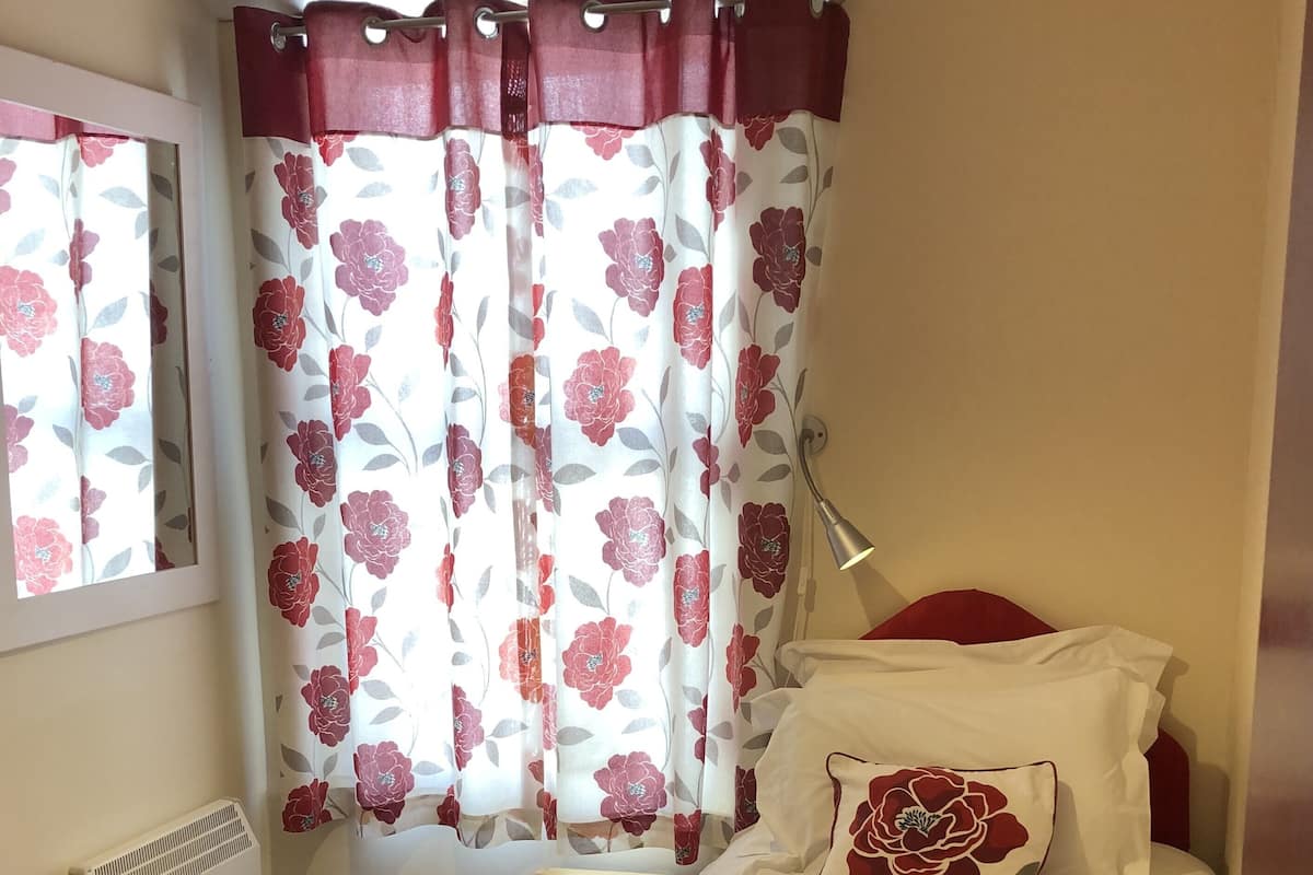 Ett sovrum med en säng, rött och vitt sänglinne, en röd pläd, en kudde med ett blommönster, ett sängbord, en lampa, ett element och gardiner med ett blommönster.