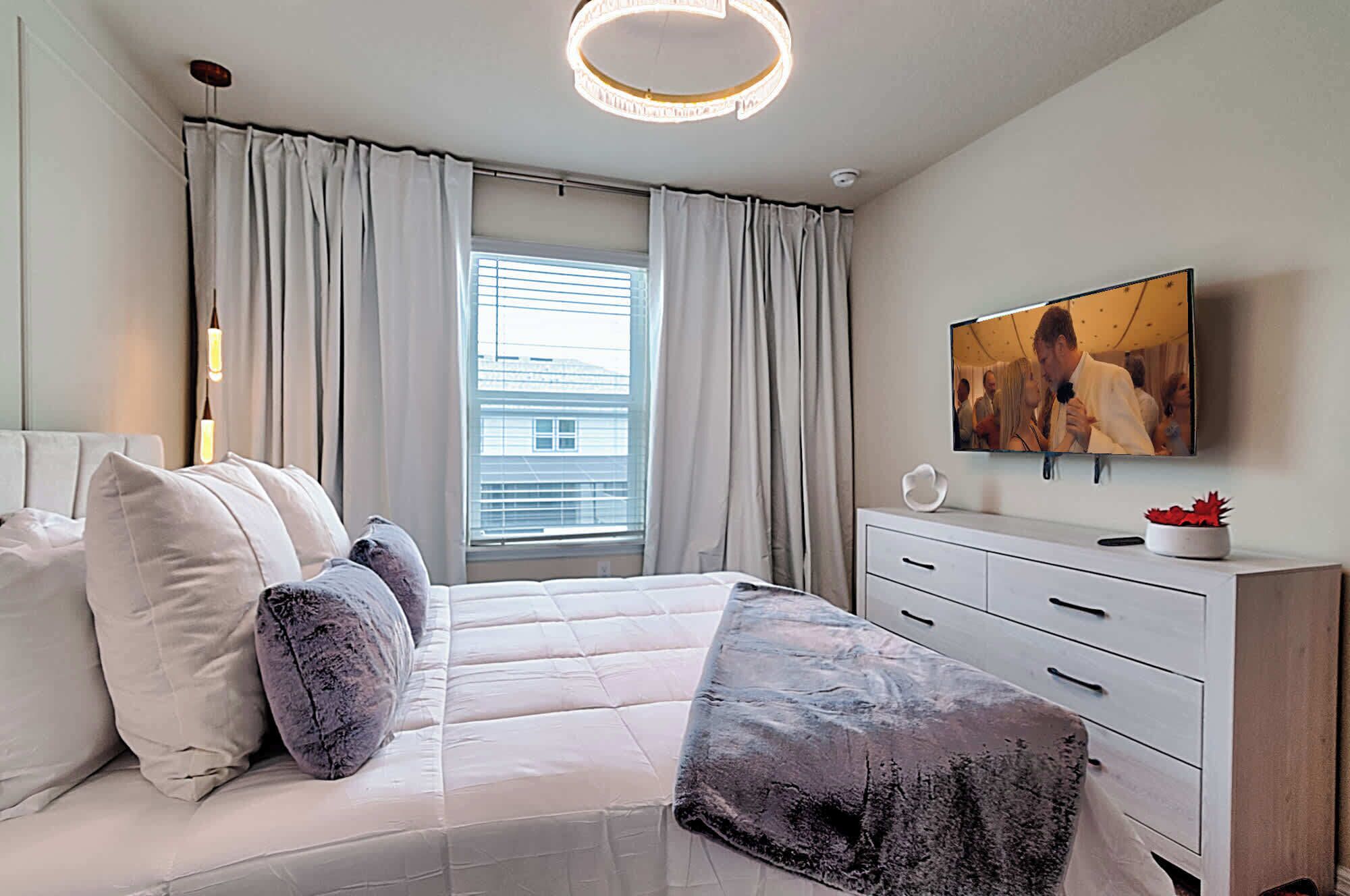 Une chambre moderne avec un lit, un téléviseur fixé au mur, une fenêtre avec des rideaux et une table de chevet agrémentée d’une plante.