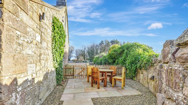 A stone wall with ivy, a wooden table and chairs, and a gravel pathway.