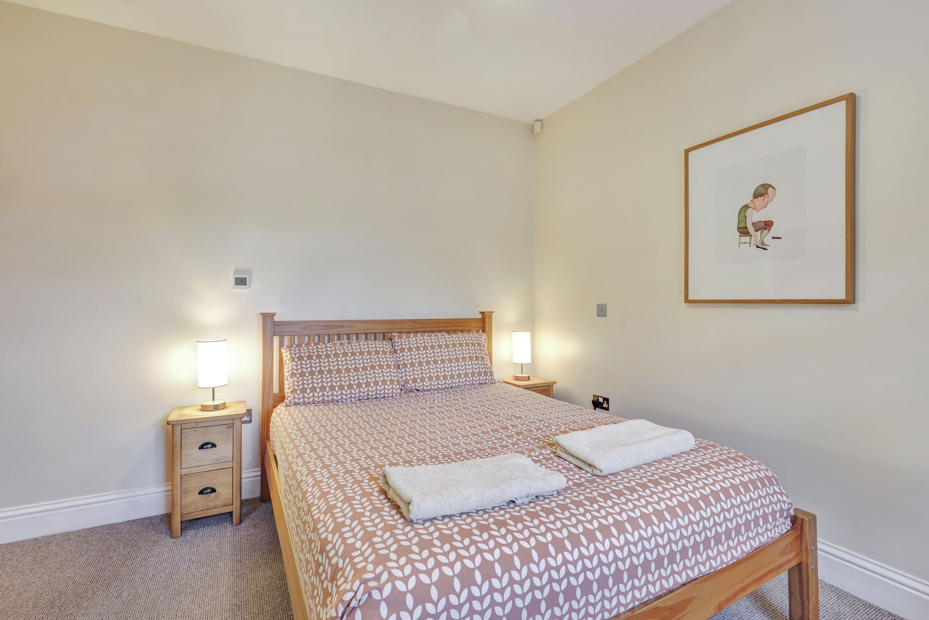 A bedroom with a bed, two bedside tables with lamps, a framed picture on the wall, and a carpeted floor.