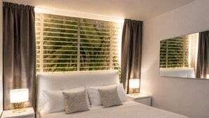A modern bedroom with a large bed, two bedside tables with lamps, a large window with blinds, and a mirror on the wall.