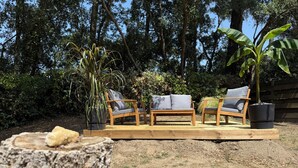 Una terraza de madera con dos sillas y un banco, rodeada de follaje y árboles.