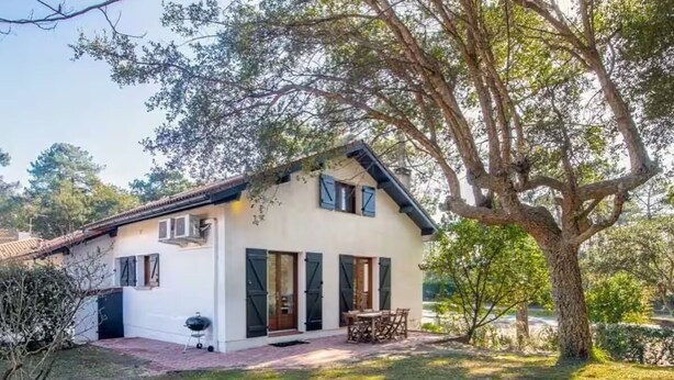Una casa de un piso con fachada blanca, un patio de ladrillo y una mesa y sillas de madera en el exterior.