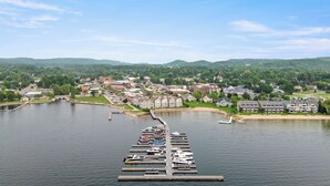 Un puerto deportivo con barcos amarrados, un puente que conecta con un pueblo con edificios residenciales y comerciales, y colinas verdes al fondo.