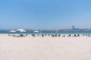 Une plage bordée de rangées de transats et de parasols, un ciel dégagé et, au loin, le profil d’une ville.