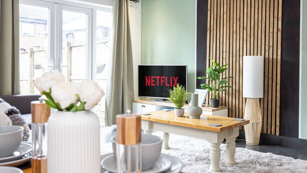 A modern living room with a wooden coffee table, white decorative bowls, a vase with white flowers, a television displaying 'NETFLIX', and a potted plant. - Relaxing 4BR Near City Centre  (Salford)