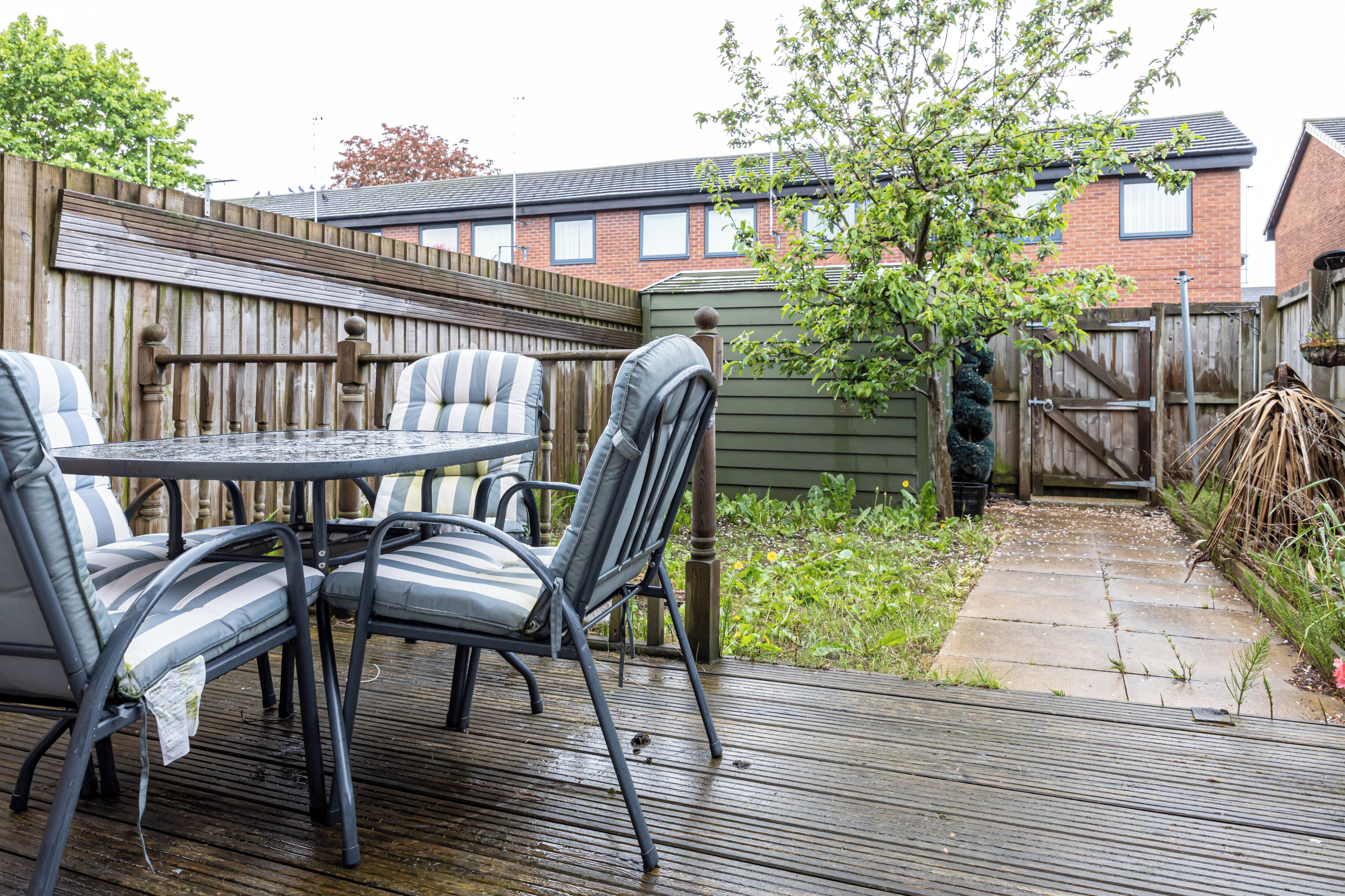 A patio with a round table and chairs, a wooden deck, and a brick house in the background.