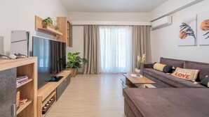 A modern living room with a flat-screen TV, wooden shelving, a sofa with decorative pillows, and a large window with curtains. - Residence City Retreat (Rodos)