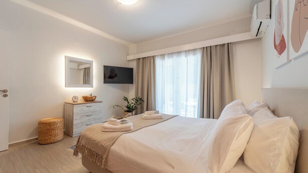 A modern hotel room with a large bed, a flat-screen TV, a bedside table with a lamp, and a window with curtains. - Residence City Retreat (Rodos)
