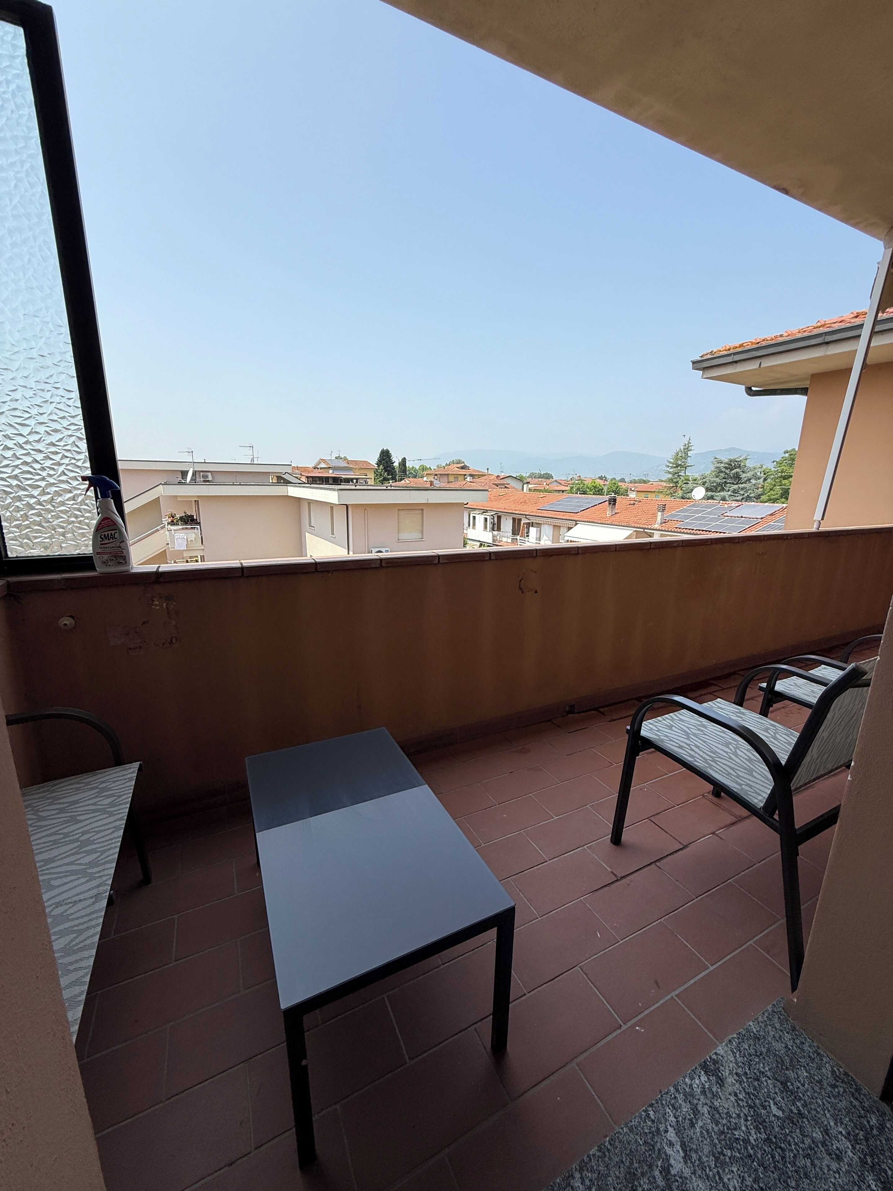 Un balcon avec une table et des chaises, une bouteille de produit nettoyant et une vue sur des immeubles et des montagnes.