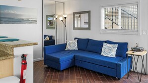 Ein modernes Wohnzimmer mit einem blauen Ecksofa, einem kleinen Beistelltisch, einem großen Spiegel und einem Fenster mit Jalousien.