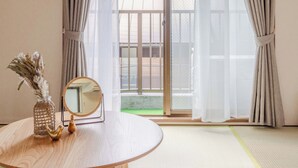 A room with tatami flooring, a round wooden table with a mirror, a vase with dried flowers, and a view through a sliding glass door to an outdoor area.