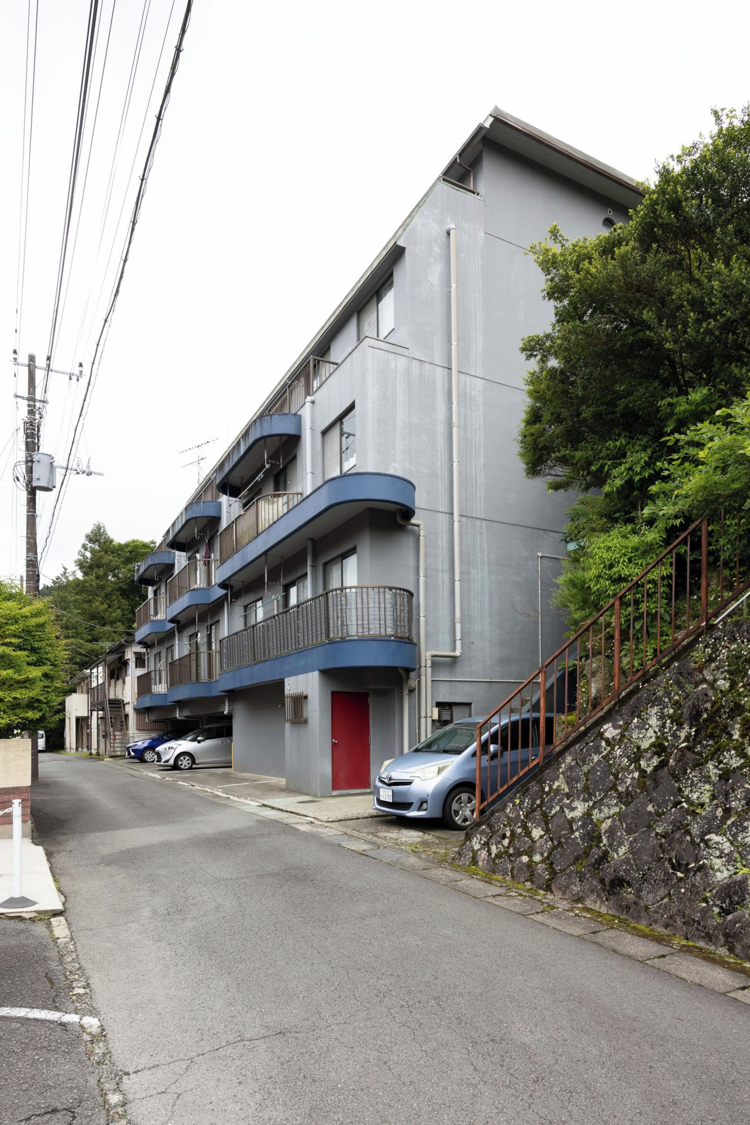 A multi-story residential building with balconies, a red door, and a staircase leading to the entrance.