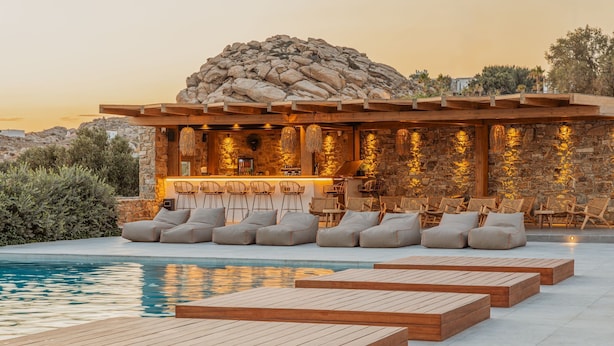Un área junto a la piscina con mobiliario de cubierta de madera y una zona de bar con paredes de piedra y luces colgantes.