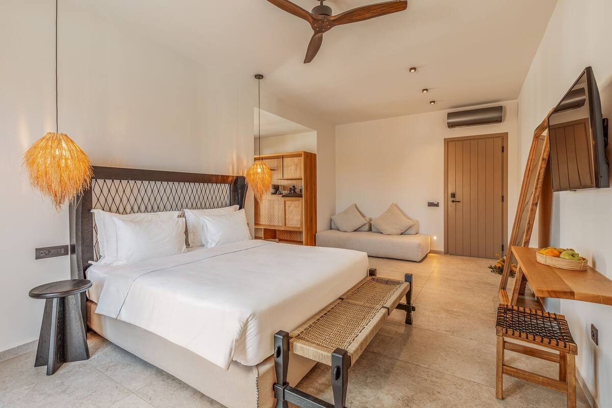 Un dormitorio moderno con una cama grande, un cabecero de madera, una mesita de noche trenzada, un banco, un sofá y un escritorio de madera con una cesta de fruta.