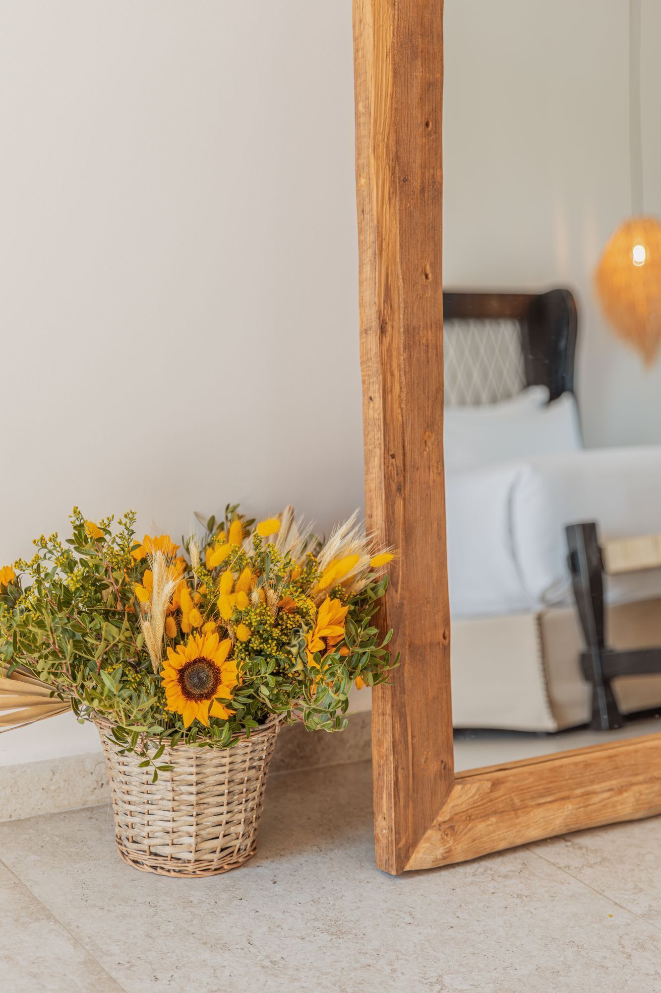 A wicker basket with sunflowers and wheat stalks on a tiled floor next to a wooden-framed mirror.