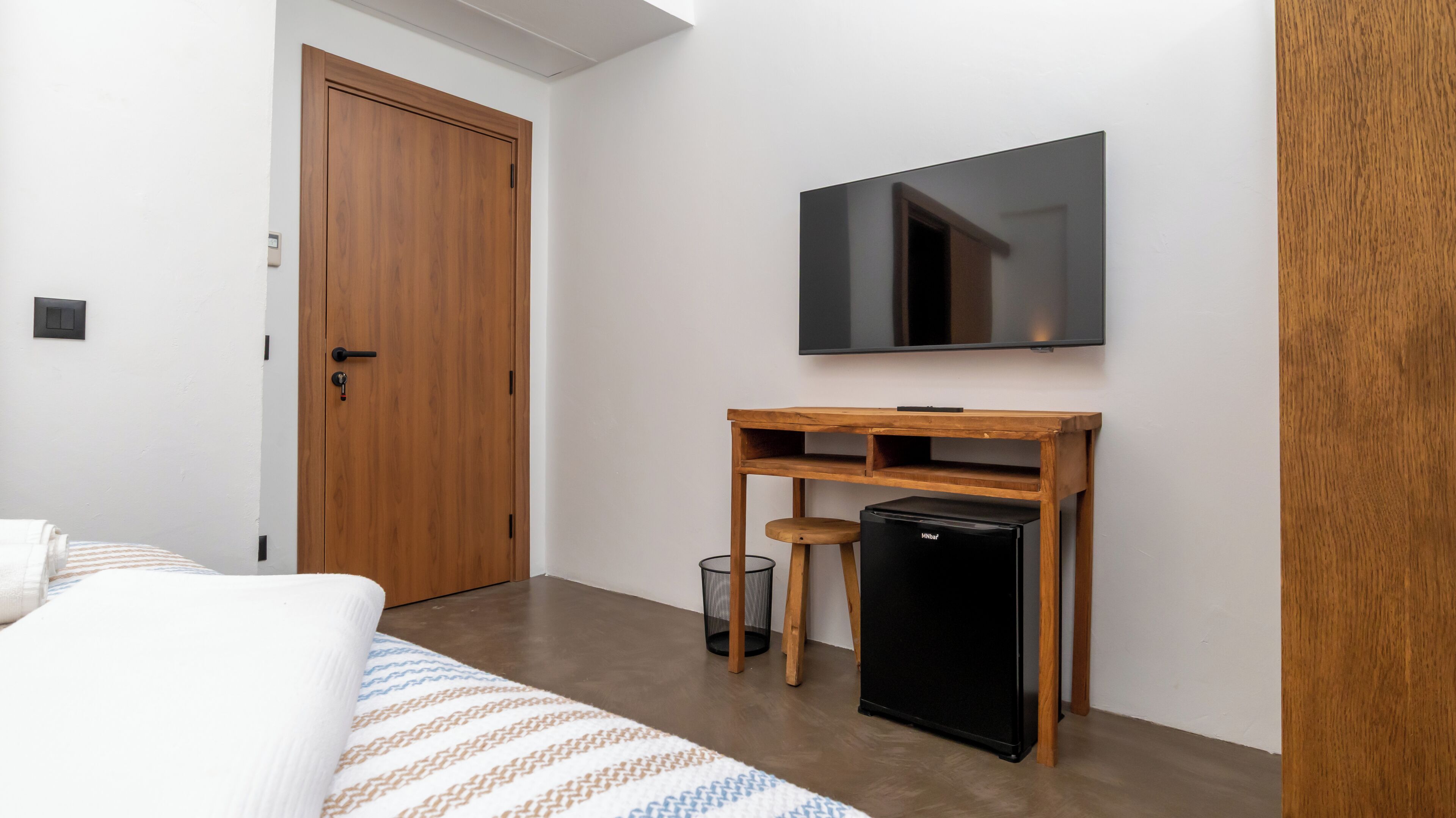 A bedroom with a bed, a wooden desk with a television, a small refrigerator, and a door.