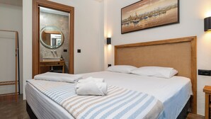 Habitación de hotel con una cama grande, cabecera de madera, un espejo redondo, un cuadro de un puerto y una mesita de noche con lámpara.