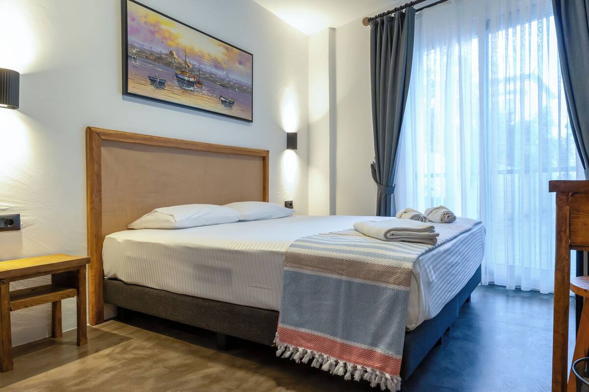 A hotel room with a bed, a desk, a chair, a painting on the wall, and a window with curtains.