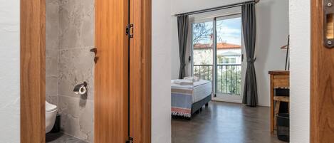 A bedroom with a bed, a window with curtains, a balcony, and a wooden door.
