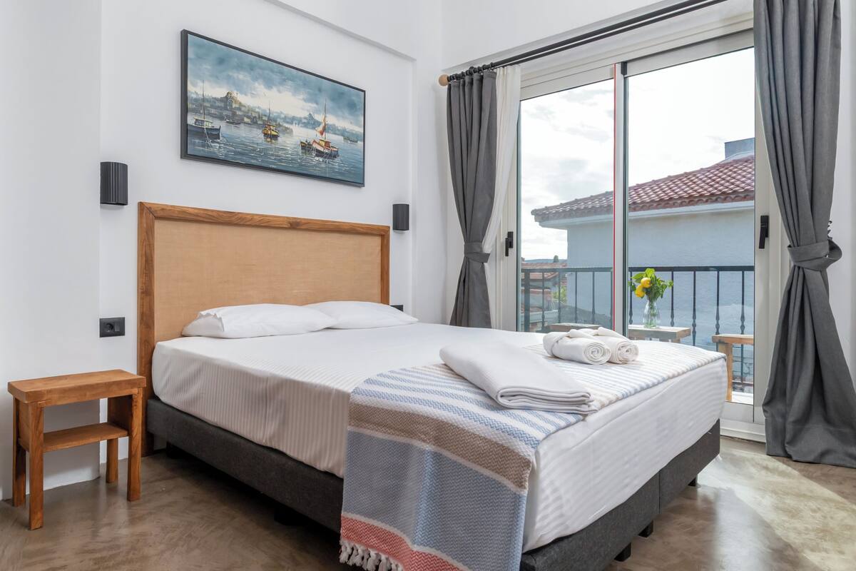 A hotel room with a bed, a small wooden nightstand, a painting of a harbor scene, and a view of a balcony with a railing.