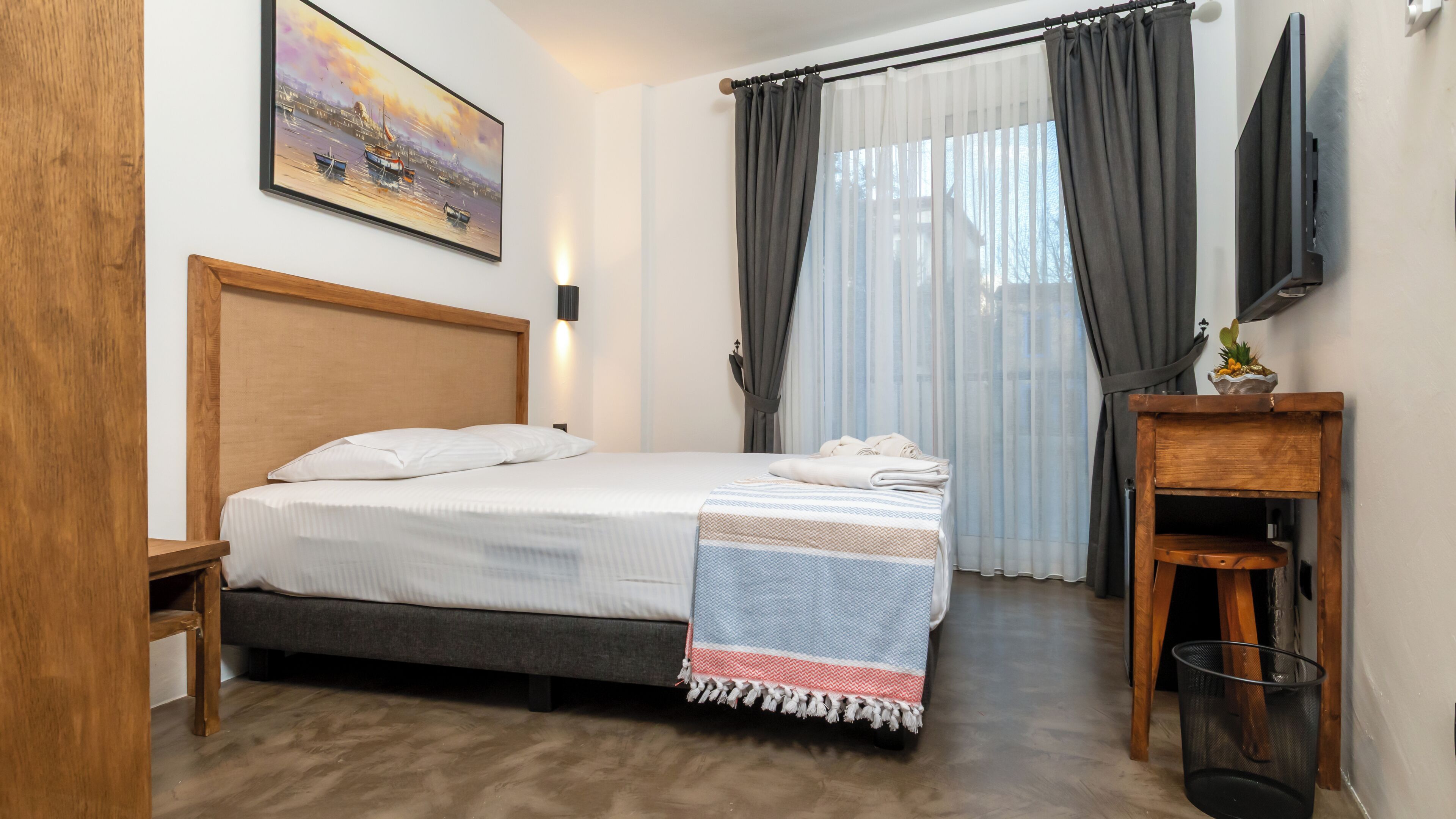 A neatly arranged bedroom with a bed, a bedside table, a small wooden nightstand, a TV mounted on the wall, and a painting above the bed.