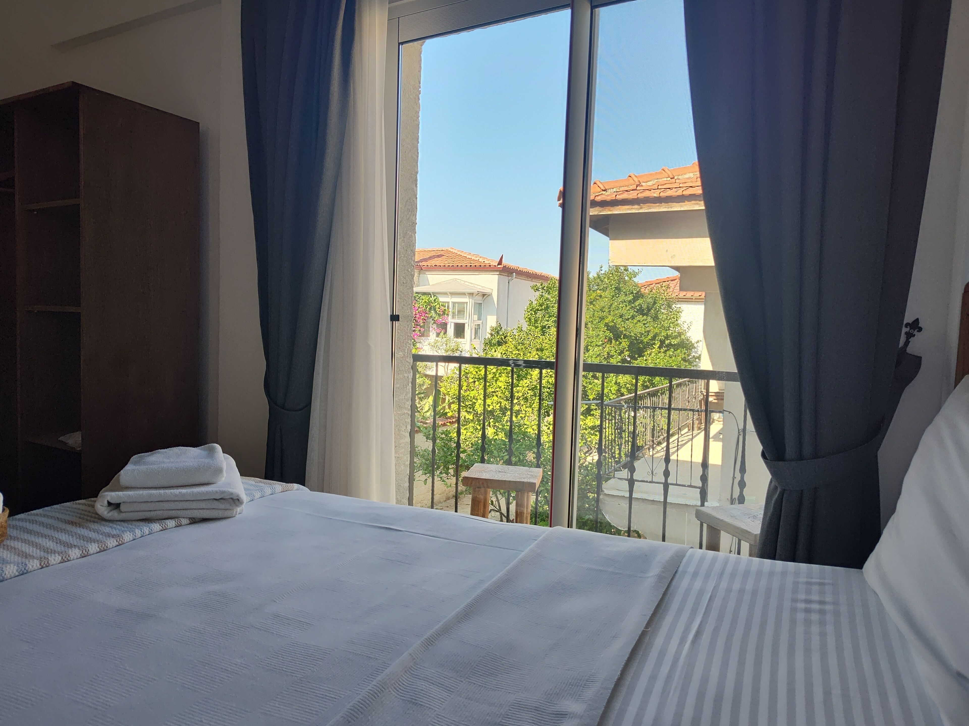 Habitación de hotel con una cama grande, un armario de madera y un balcón con vistas a edificios y árboles.