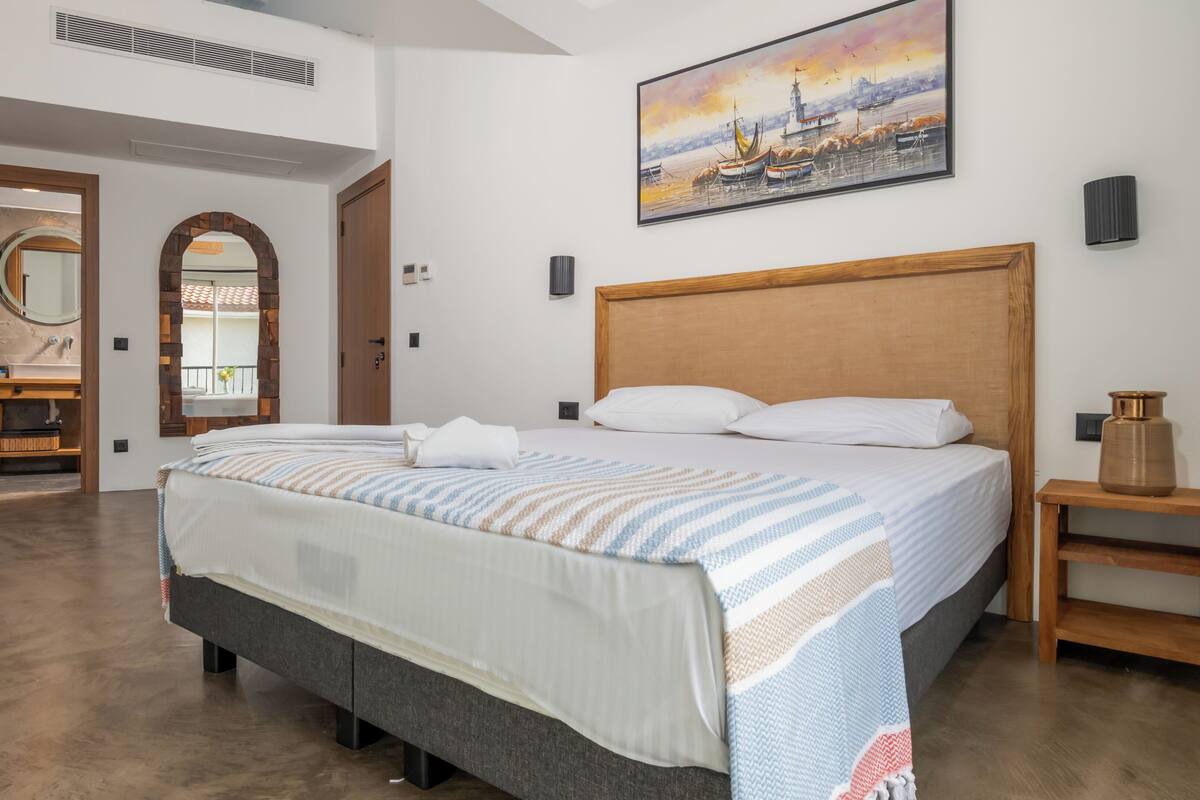 A hotel room with a large bed, a painting on the wall, and a view through an arched window.
