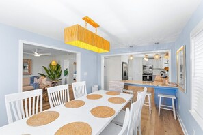 Un comedor con una mesa y sillas blancas, una lámpara colgante y una cocina al fondo.