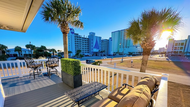 A deck with outdoor furniture, a planter, and a view of a cityscape with buildings and palm trees. - The Beach Jewel - 100% Newly Renovated in 2024! (North Myrtle Beach)